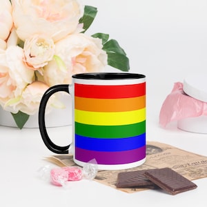 Könnte beinhalten: Eine Keramik-Tasse mit schwarzem Henkel und Rand, die ein Regenbogen-Design mit horizontalen Streifen in Rot, Orange, Gelb, Grün, Blau und Lila aufweist. Die Tasse steht vor Blumen und Schokolade.