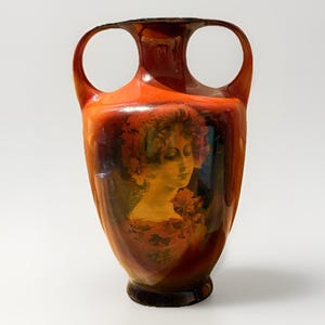 Op de afbeelding: Een decoratieve, oranjerode vaas met twee handvatten en een portret van een vrouw. De vaas heeft een donkerbruine basis en rand. Het portret van de vrouw is in tinten geel, oranje en bruin.