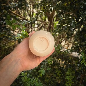 May include: A small, round, off-white ceramic bowl held in a hand. The bowl has a smooth exterior and a slightly textured interior. The bottom of the bowl features a small, stamped design. The background is a blurred view of green foliage.