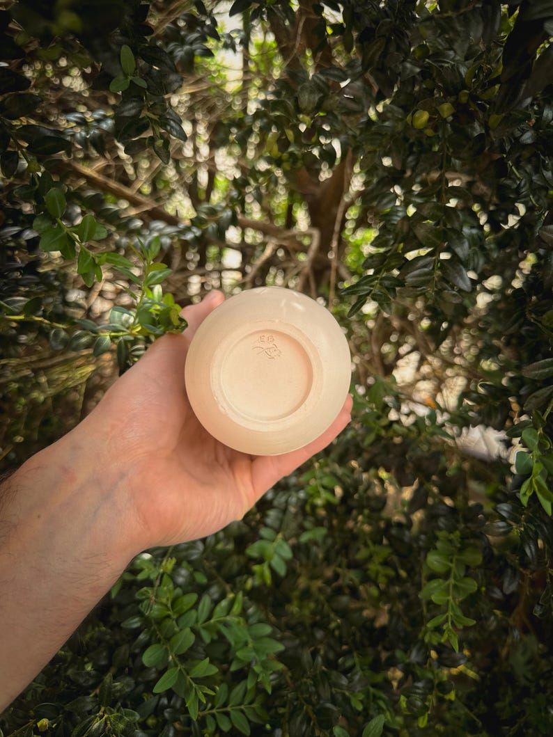 May include: A small, round, off-white ceramic bowl held in a hand. The bowl has a smooth exterior and a slightly textured interior. The bottom of the bowl features a small, stamped design. The background is a blurred view of green foliage.
