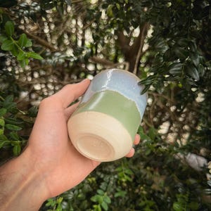 May include: A handmade ceramic tumbler with a unique color-blocked design. The top section is a light blue, the middle is green, and the base is a natural beige. The tumbler is held in a hand, showcasing its smooth, glazed finish.