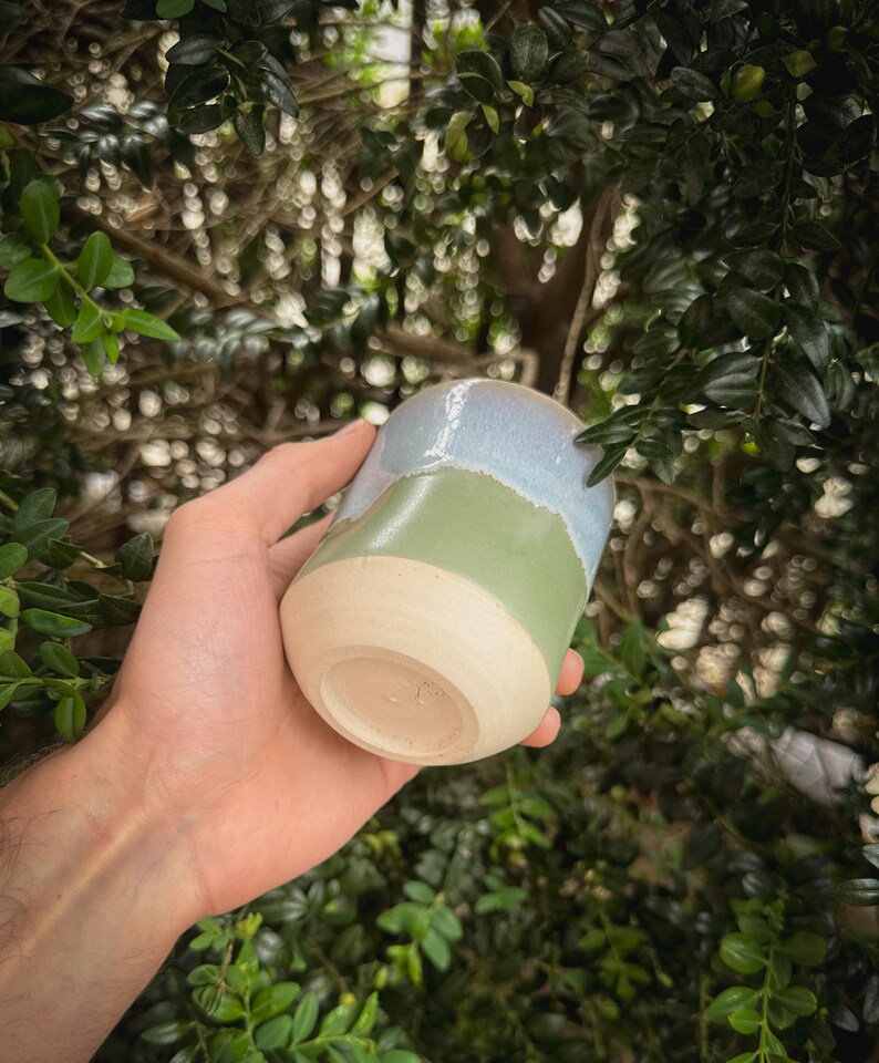 May include: A handmade ceramic tumbler with a unique color-blocked design. The top section is a light blue, the middle is green, and the base is a natural beige. The tumbler is held in a hand, showcasing its smooth, glazed finish.
