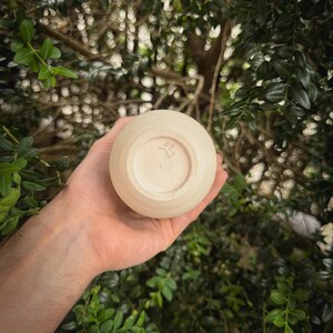 May include: A small, round, beige ceramic cup with a stamped design on the bottom. The cup is held in a hand, with a blurred green leafy background. The cup appears to be handmade.