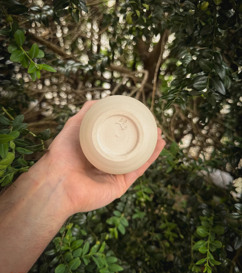 May include: A small, round, beige ceramic cup with a stamped design on the bottom. The cup is held in a hand, with a blurred green leafy background. The cup appears to be handmade.