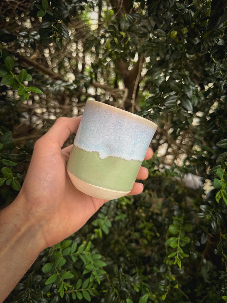 May include: A handmade ceramic mug with a light blue top, a green middle section, and a beige base. The mug is held against a backdrop of green foliage, showcasing its unique color scheme and artisanal quality.