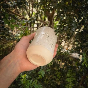 May include: A handmade ceramic tumbler with a speckled white glaze and a natural, unglazed base. The cylindrical cup is held against a backdrop of green foliage, showcasing its handcrafted quality and organic aesthetic.
