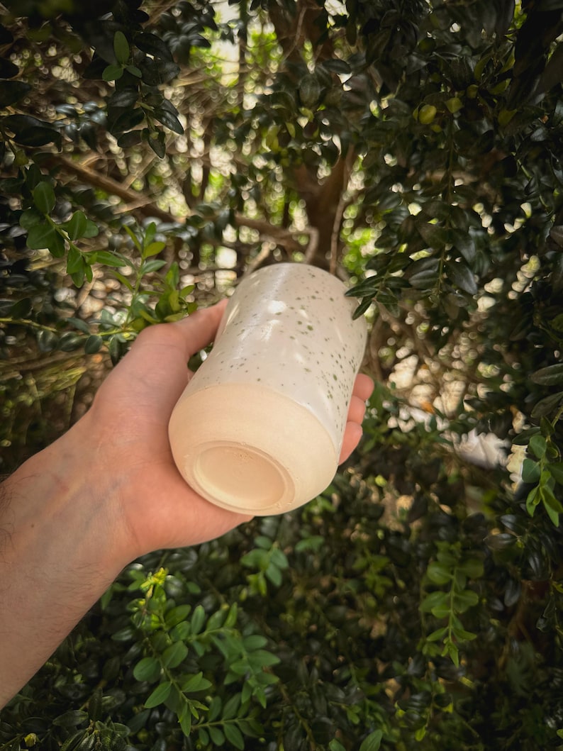 May include: A handmade ceramic tumbler with a speckled white glaze and a natural, unglazed base. The cylindrical cup is held against a backdrop of green foliage, showcasing its handcrafted quality and organic aesthetic.