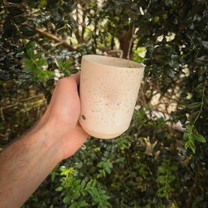 May include: A speckled ceramic mug in a light beige color is held against a backdrop of green foliage. The mug has a cylindrical shape with a slightly flared rim and a smooth, unglazed base. The surface is dotted with small, dark specks.