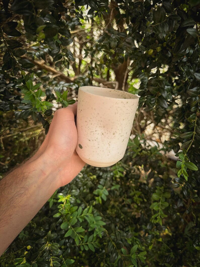 May include: A speckled ceramic mug in a light beige color is held against a backdrop of green foliage. The mug has a cylindrical shape with a slightly flared rim and a smooth, unglazed base. The surface is dotted with small, dark specks.
