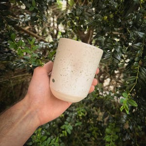 May include: A speckled ceramic mug with a light beige base and a cylindrical shape. The mug has a speckled pattern of small green dots and a smooth, rounded bottom. The mug is held in a hand against a backdrop of green foliage.