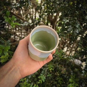 May include: A handmade ceramic cup with a green interior and a light blue and beige exterior. The cup is held in a hand against a backdrop of green foliage. The cup's design is simple and elegant, suitable for beverages.