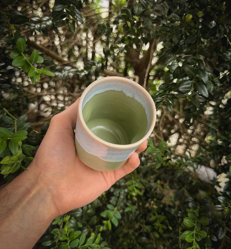 May include: A handmade ceramic cup with a green interior and a light blue and beige exterior. The cup is held in a hand against a backdrop of green foliage. The cup's design is simple and elegant, suitable for beverages.