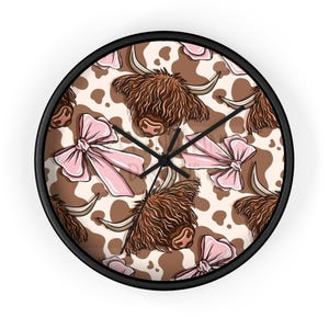 May include: A round wall clock with a black frame. The clock face features a repeating pattern of brown Highland cow faces, pink bows, and a cowhide design. The clock hands are black.