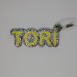 Pode incluir: Um porta-chaves com o nome "TORI" em letras amarelo brilhante, contornado a preto. As letras são incrustadas com strass iridescentes. Uma argola prateada está presa no canto superior direito.