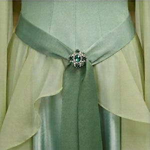 Puede incluir: Primer plano de un vestido verde claro con una faja decorativa. La faja es de un tono verde m&aacute;s oscuro y est&aacute; adornada con un broche brillante con una gran piedra verde ovalada y piedras claras m&aacute;s peque&ntilde;as. El vestido tiene un dise&ntilde;o fluido y en capas.