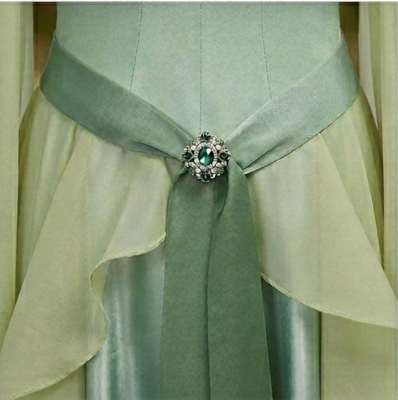 Puede incluir: Primer plano de un vestido verde claro con una faja decorativa. La faja es de un tono verde m&aacute;s oscuro y est&aacute; adornada con un broche brillante con una gran piedra verde ovalada y piedras claras m&aacute;s peque&ntilde;as. El vestido tiene un dise&ntilde;o fluido y en capas.