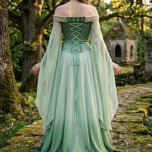 Puede incluir: Un vestido largo y fluido de color verde con un corpi&ntilde;o de cors&eacute; y mangas con hombros descubiertos. El vestido presenta un efecto degradado, pasando de un verde m&aacute;s oscuro en el corpi&ntilde;o a un tono m&aacute;s claro en la falda. Las mangas son largas y vaporosas.