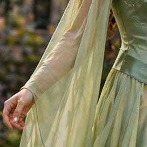 Puede incluir: Un vestido verde claro con mangas largas y fluidas. El vestido presenta un corpi&ntilde;o ajustado y una falda en capas. Las mangas son de tela transparente y el vestido tiene un ribete dorado alrededor del escote. Adecuado para una feria renacentista o cosplay.