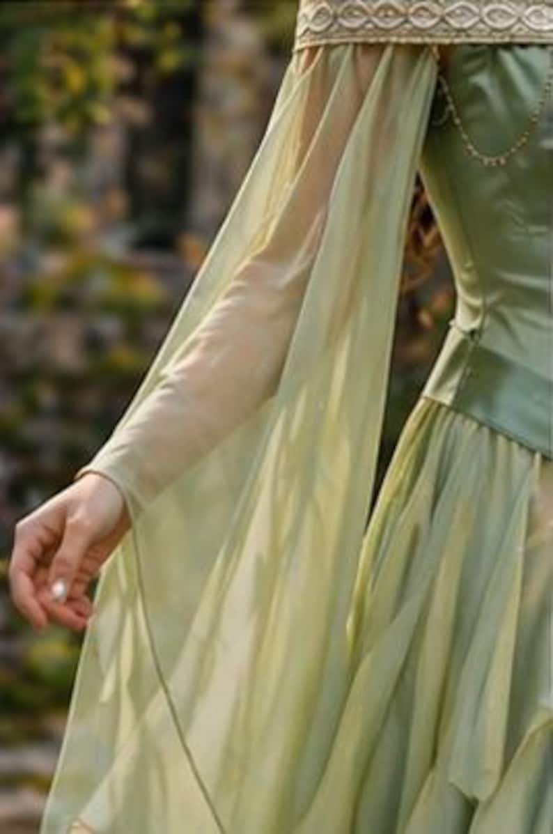 Puede incluir: Un vestido verde claro con mangas largas y fluidas. El vestido presenta un corpi&ntilde;o ajustado y una falda en capas. Las mangas son de tela transparente y el vestido tiene un ribete dorado alrededor del escote. Adecuado para una feria renacentista o cosplay.