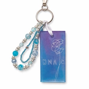 Puede incluir: Un llavero con un dije rectangular con un fondo degradado azul y morado. El dije tiene la palabra "DNA" y un diseño floral en blanco. El llavero incluye una correa de muñeca con cuentas azules y blancas y un cierre plateado.