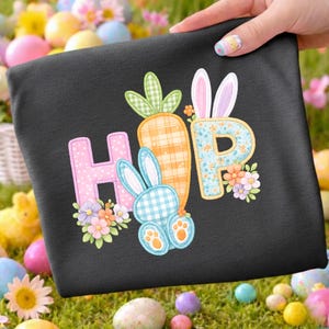 May include: Black t-shirt with a colorful "HOP" design featuring a carrot, bunny, and flowers. The letters are decorated with patterns and surrounded by floral accents, perfect for Easter celebrations.