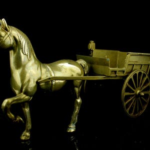 May include: A brass horse figurine pulling a small cart. The horse is in a trotting pose, with one leg raised. The cart has spoked wheels and a rectangular bed. The entire piece has a shiny, metallic finish.