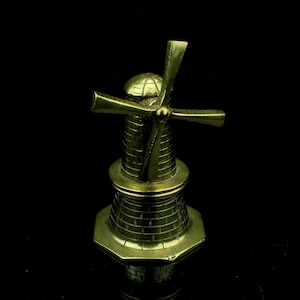 Puede incluir: Una figura de molino de viento de latón con un diseño detallado. El molino tiene un cuerpo cilíndrico con un patrón de ladrillos y una base hexagonal. Las aspas están colocadas en la parte superior. La figura está sobre un fondo negro.