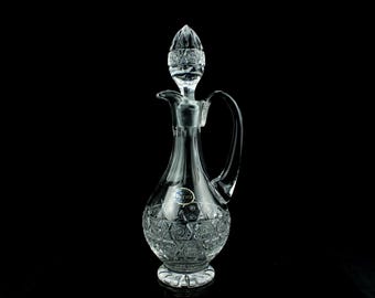May include: A clear crystal decanter featuring intricate geometric cut patterns on the base and a matching pointed stopper. The body has a curved handle and a spout, with a small blue and gold label reading BOHEMIA attached to the front surface.
