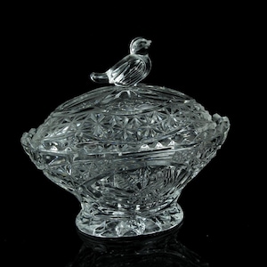 Può includere: Un portagioie in vetro tagliato trasparente con coperchio, con una dettagliata statuina di uccello sulla parte superiore. Il piatto presenta un motivo decorativo e strutturato e poggia su una piccola base a piedistallo. Il design generale è elegante e vintage.