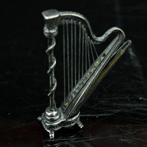 Op de afbeelding: Een gedetailleerde zilverkleurige miniatuurharp met snaren, een decoratieve spiraalvormige kolom en een sierlijke basis. De harp staat tegen een donkere, reflecterende achtergrond, wat het ingewikkelde ontwerp en vakmanschap benadrukt. De snaren van het instrument zijn zichtbaar.