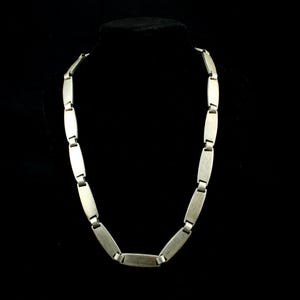 May include: A silver-toned necklace composed of a series of rectangular, brushed-finish metal links connected by small, rounded hinges. The chain features a uniform design with a matte texture, creating a continuous, structured loop of metal segments.