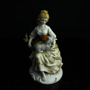 Peut inclure: Figurine en porcelaine d'une femme en robe longue tenant un agneau. La robe de la femme est crème avec des motifs floraux rouges. Elle a les cheveux blonds et est assise sur une base blanche avec une bordure dorée. Le fond est noir.