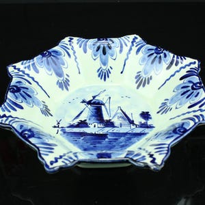 Pode incluir: Uma tigela de cerâmica decorativa com borda ondulada e uma cena de moinho de vento azul e branco. A tigela apresenta uma pintura detalhada da paisagem de um moinho de vento, casas e água. A borda é adornada com padrões florais.