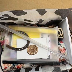 May include: A white gift box lined with black and white cow-print tissue paper containing a bronze-colored metal horseshoe, a circular wax seal with a horseshoe emblem, a white card, and a floral-patterned cylindrical object in a plastic bag.