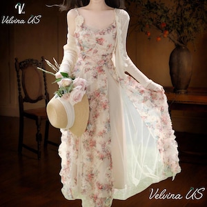 May include: A floral print dress with a matching long-sleeve cardigan. The dress features a V-neck and a flowing skirt. A straw hat with a white ribbon and pink roses is held in the hand. The text "Velvina US" is in the upper left and lower right corners.