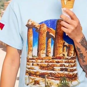 Puede incluir: Una camiseta de manga corta de color azul claro con un estampado gráfico de columnas de piedra antiguas sobre un cielo azul. La prenda incluye un pequeño emblema rectangular en la manga derecha con un diseño rojo y amarillo.
