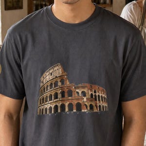 May include: A charcoal gray short-sleeve t-shirt featuring a detailed graphic print of the Roman Colosseum on the front center and a small circular emblem on the right sleeve. The shirt has a crew neckline and a relaxed fit, constructed from cotton.
