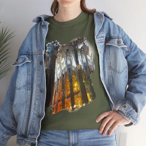 May include: An olive green crew neck t-shirt featuring a graphic print of a cathedral interior with tall, dark, branching columns and warm, glowing light. The shirt is worn under a light blue denim jacket with silver buttons and visible stitching.