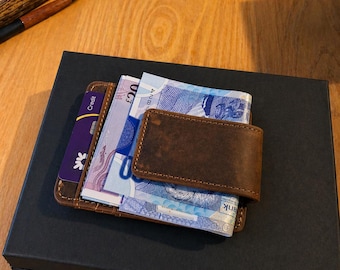 Könnte beinhalten: Ein braunes Lederportemonnaie mit einem Geldscheinclip, das mehrere Geldscheine und eine Kreditkarte enthält. Das Portemonnaie liegt auf einer schwarzen Box, was auf ein Geschenk oder einen Aufbewahrungsgegenstand hindeutet. Der Gesamteindruck ist hochwertig.