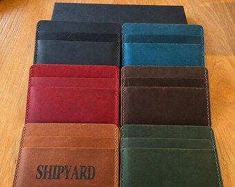 Könnte beinhalten: Eine Sammlung von sechs rechteckigen Kartenetuis aus Leder, die in zwei Spalten angeordnet sind. Die Farben umfassen Schwarz, Blaugrün, Rot, Dunkelbraun, Hellbraun und Waldgrün. Das hellbraune Kartenetui trägt das Wort SHIPYARD in schwarzer Schrift.