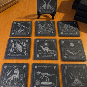 Puede incluir: Un juego de diez posavasos cuadrados de pizarra con ilustraciones de dinosaurios grabadas con l&aacute;ser al estilo de cartas de tarot. Cada posavasos muestra una criatura prehist&oacute;rica, n&uacute;meros romanos y t&iacute;tulos como The Lovers o The Tower.