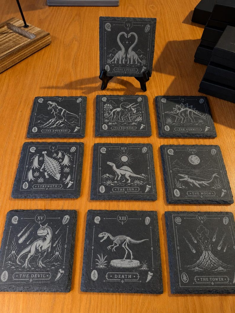Puede incluir: Un juego de diez posavasos cuadrados de pizarra con ilustraciones de dinosaurios grabadas con l&aacute;ser al estilo de cartas de tarot. Cada posavasos muestra una criatura prehist&oacute;rica, n&uacute;meros romanos y t&iacute;tulos como The Lovers o The Tower.