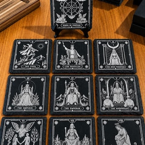 May include: A set of square black slate coasters featuring white engraved tarot card illustrations. The designs include The Wheel of Fortune, The Fool, The Magician, The High Priestess, The Empress, The Emperor, The Hierophant, The Lovers, The Chariot, and Strength.