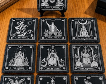 Könnte beinhalten: Ein Set quadratischer Untersetzer aus schwarzem Schiefer mit weiß gravierten Tarotkarten-Illustrationen. Die Motive zeigen das Rad des Schicksals, den Narren, den Magier, die Hohepriesterin, die Herrscherin, den Herrscher, den Hierophanten, die Liebenden, den Wagen und die Kraft.