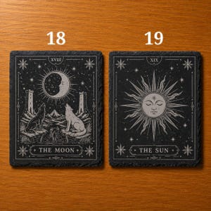 May include: Two rectangular black slate coasters featuring white engraved designs. The left coaster, labeled 18, depicts The Moon with wolves, towers, and a crustacean. The right coaster, labeled 19, depicts The Sun with a central face and rays.