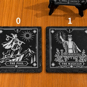 May include: Two square black slate coasters featuring white engraved tarot card designs. The left coaster displays The Fool with a figure, dog, and sun. The right coaster shows The Magician with a figure, table, and symbols. Both have white borders.