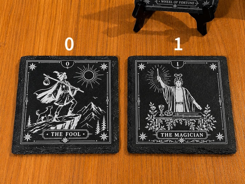 May include: Two square black slate coasters featuring white engraved tarot card designs. The left coaster displays The Fool with a figure, dog, and sun. The right coaster shows The Magician with a figure, table, and symbols. Both have white borders.
