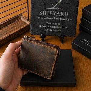 Puede incluir: Una cartera de cuero marrón con cierre de cremallera. La cartera tiene una textura desgastada y forma rectangular. En el fondo, un letrero de pizarra negra muestra el texto SHIPYARD, Local leathersmith and engraving, y ShipyardBelfast@gmail.com.