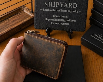 May include: A brown leather wallet with a zipper closure. The wallet has a distressed texture and a rectangular shape. In the background, a black slate sign displays the text SHIPYARD, Local leathersmith and engraving, and ShipyardBelfast@gmail.com.