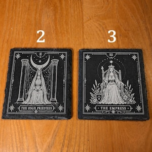 May include: Two square black slate coasters featuring white engraved tarot card designs. The left coaster displays The High Priestess with pillars and a crescent moon. The right coaster shows The Empress seated on a throne with floral details.